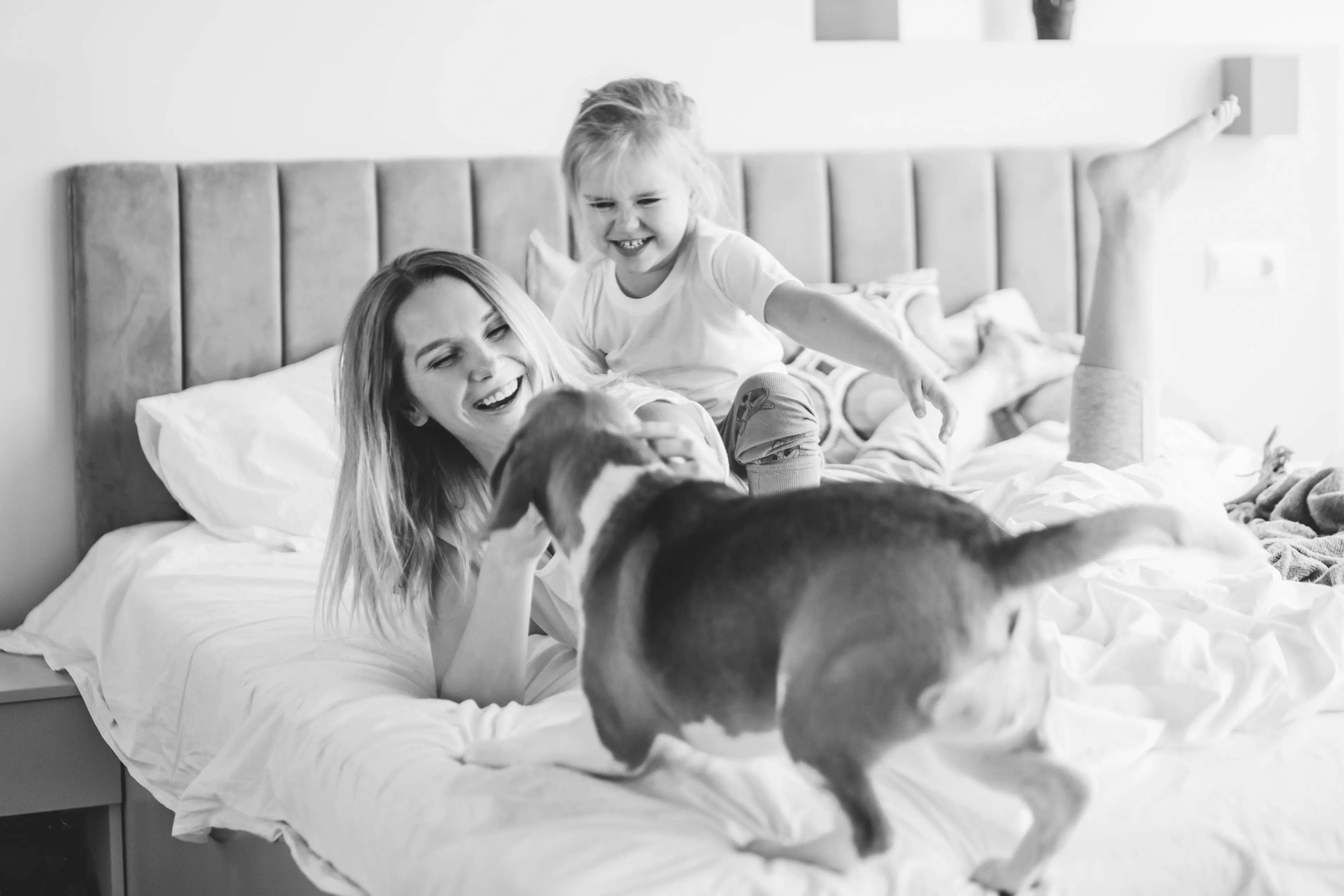 Schwarz-weiß Foto einer Mutter und ihrer Tochter, die lachend mit einem Hund auf dem Bett spielen